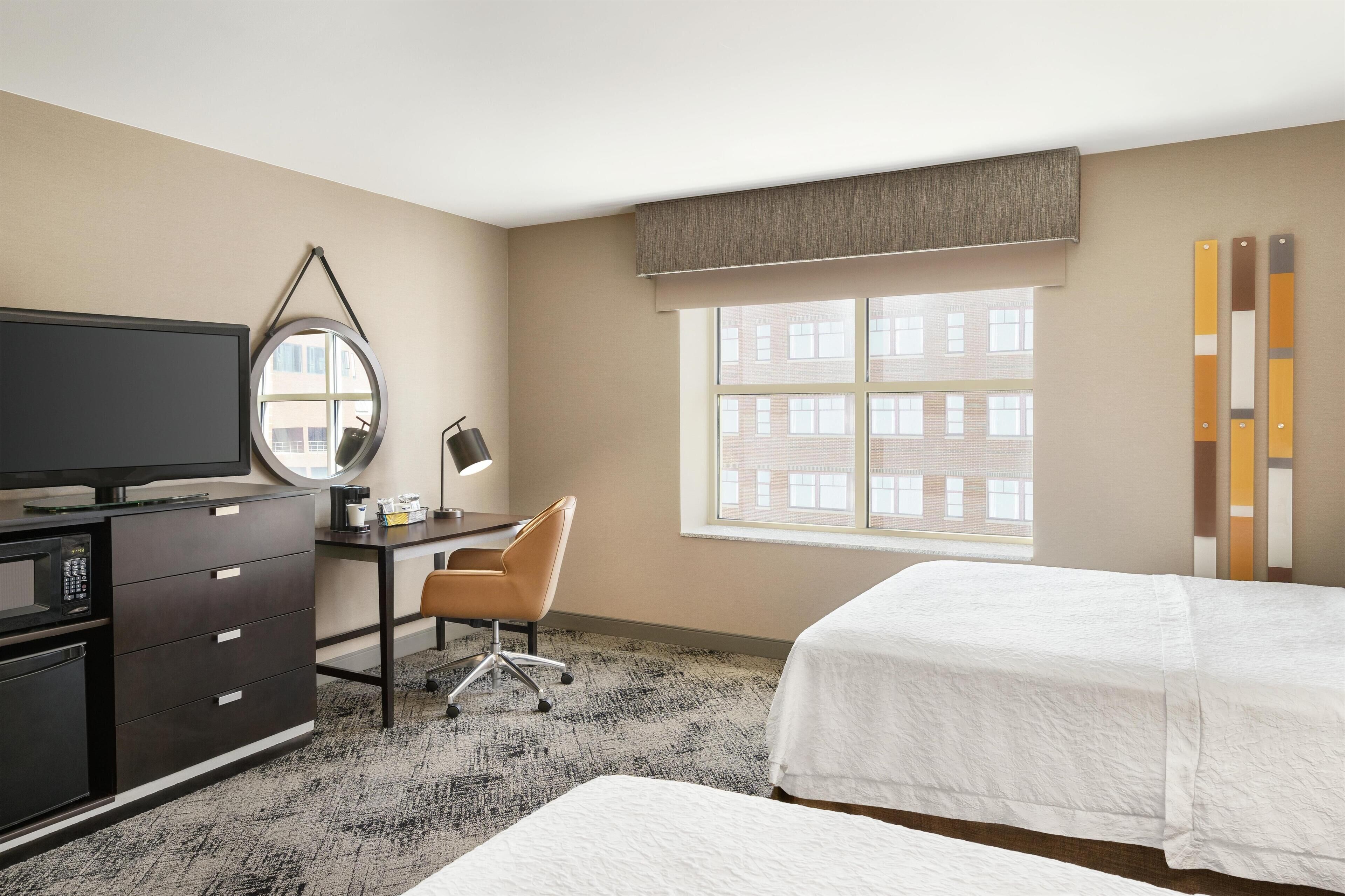 Une chambre d’hôtel comprenant un lit, un bureau avec un ordinateur, une télévision, une chaise et une fenêtre donnant sur des immeubles.