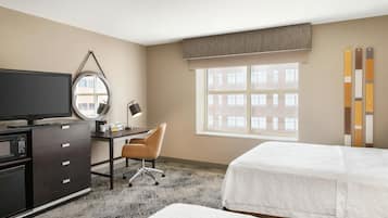 A hotel room with a bed, a desk with a computer, a television, a chair, and a window with a view of buildings.