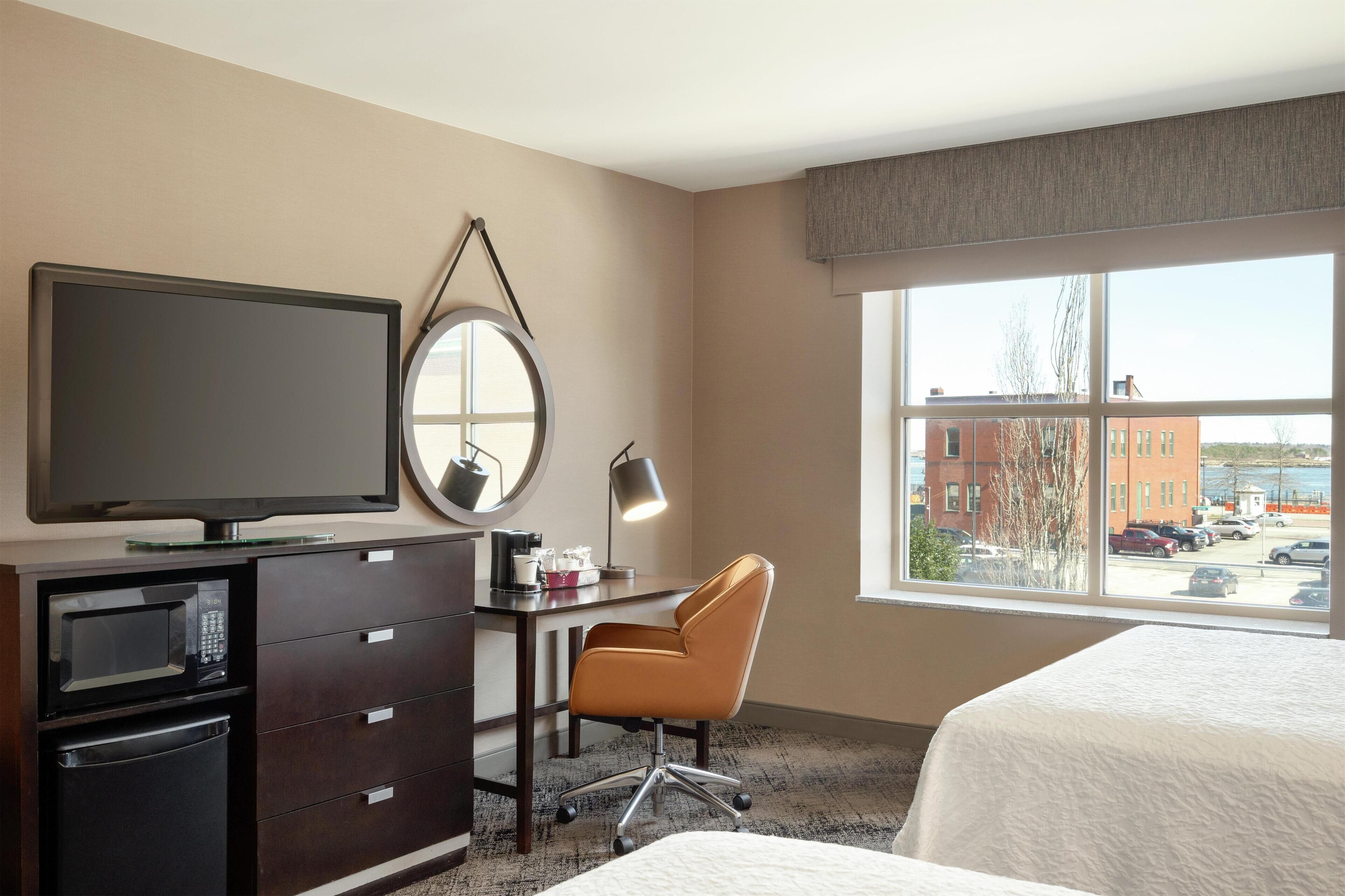 Ein Hotelzimmer mit einem Bett, einem Schreibtisch mit Stuhl, einem Fernseher, einer Mikrowelle und einem großen Fenster mit Blick auf Gebäude und Autos.
