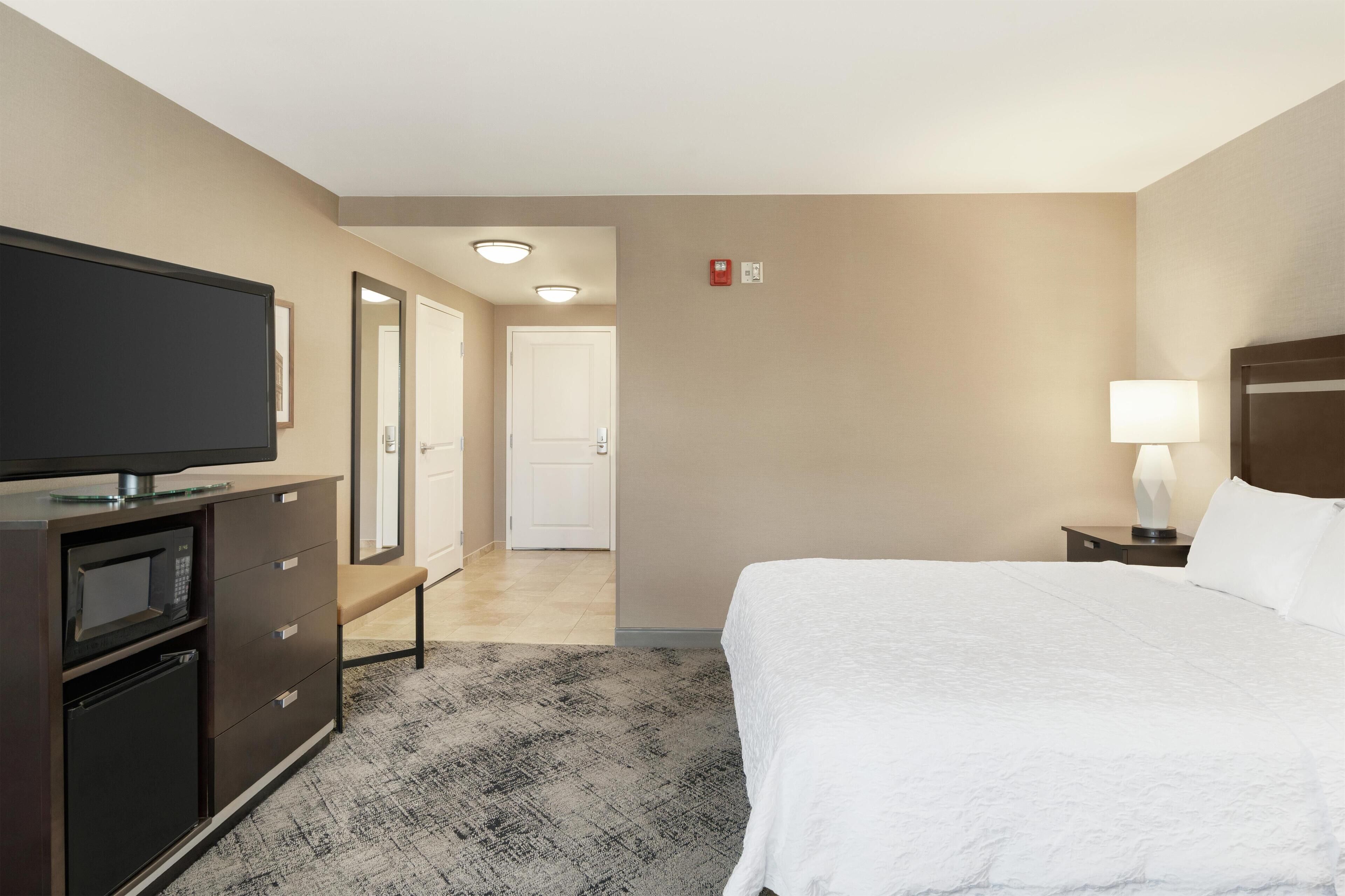 A hotel room with a bed, a TV, a desk with a lamp, a microwave, and a bench.