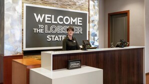 A reception desk with a "Welcome to the Lobster State" sign, a "Hilton Honors" sign, and a digital screen.