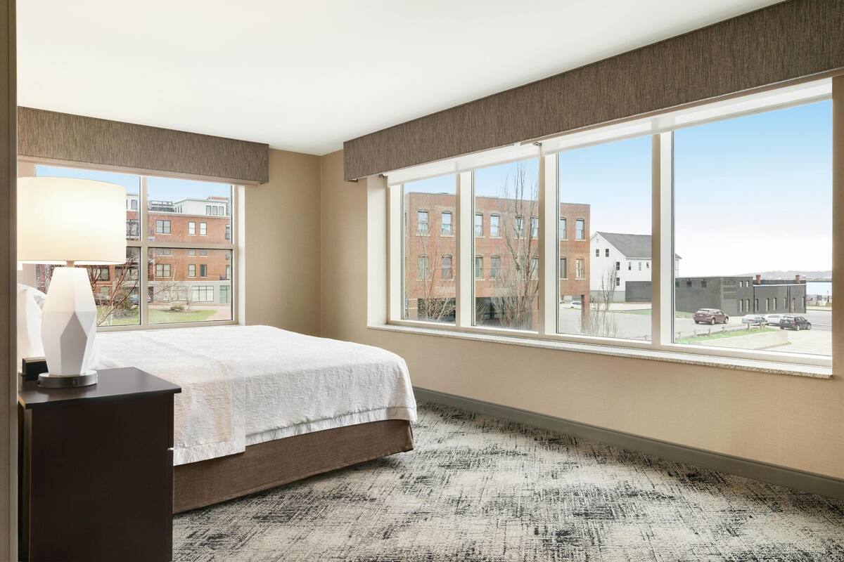 A hotel room with a large window, a bed, a nightstand, and a lamp.