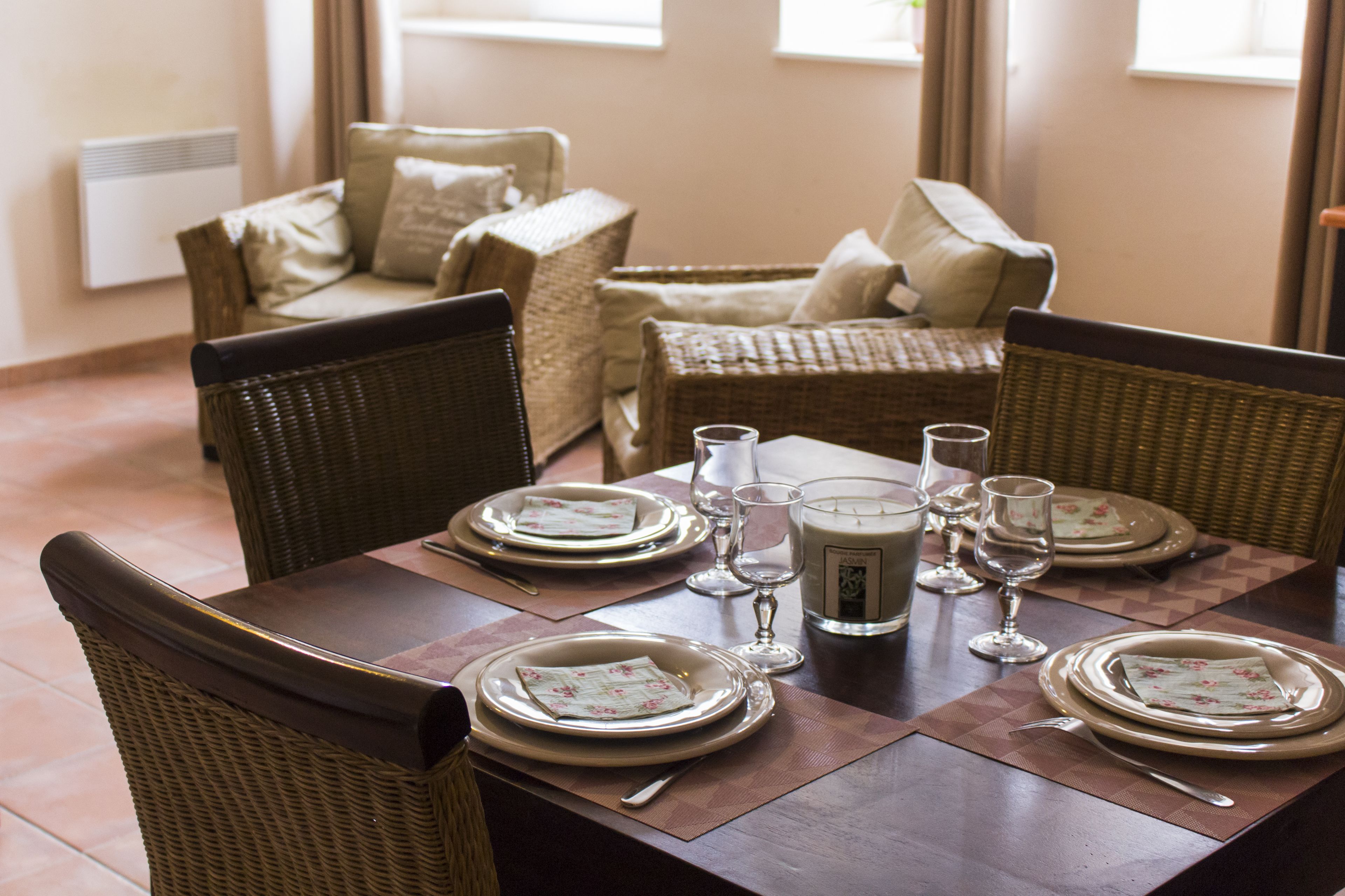 Une table à manger dressée pour quatre personnes, avec des chaises en osier, un centre de table composé de bougies et, en arrière-plan, un canapé en osier.