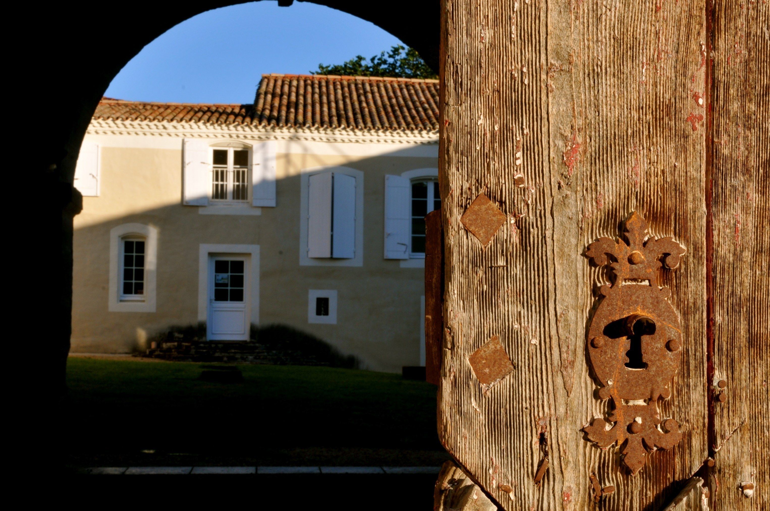 A rustic wooden door with a metal keyhole, leading to a building with a tiled roof and white shutters.