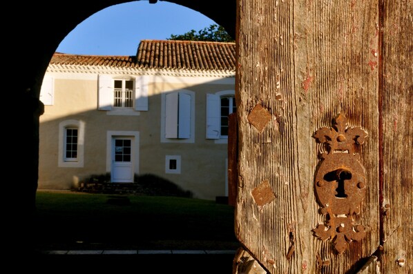 A rustic wooden door with a metal keyhole, leading to a building with a tiled roof and white shutters.