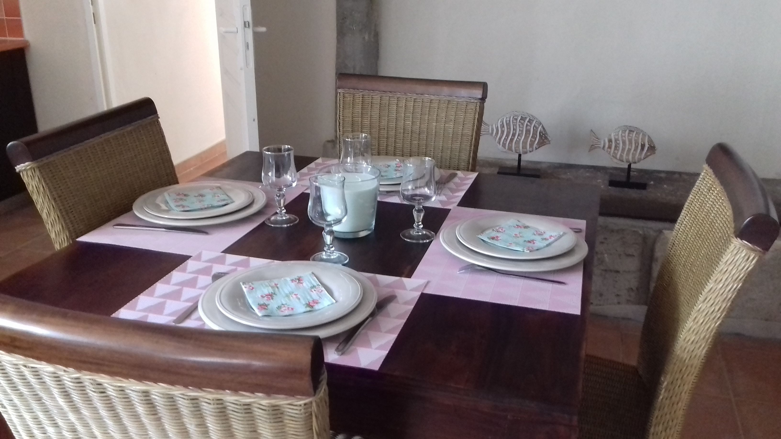 Una mesa de comedor dispuesta con platos, vasos y una jarrón, rodeada de sillas de mimbre.