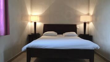 Una cama bien hecha con dos almohadas, flanqueada por dos mesitas de noche con lámparas, en una habitación con paredes lisas y suelo de baldosas.