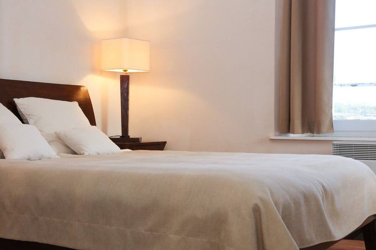Un lit bien fait, recouvert d’une couvre-lit blanche et agrémenté d’oreillers, une table de chevet avec une lampe, et une fenêtre avec des rideaux.