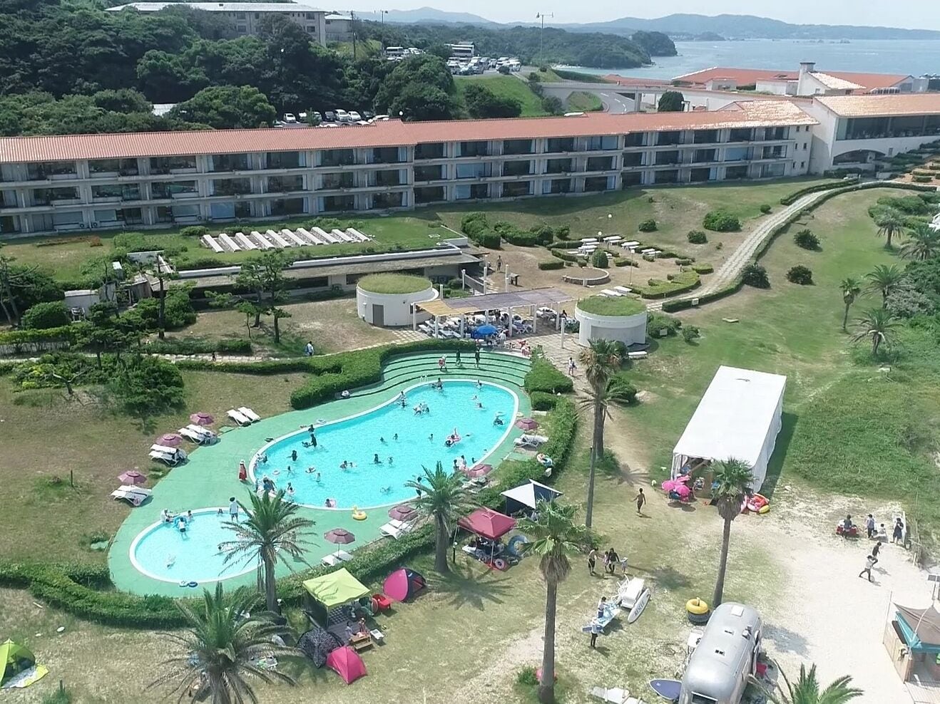 A hotel with a large swimming pool surrounded by sun loungers and umbrellas, a covered outdoor area, and a parking lot.