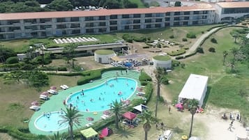 A hotel with a large swimming pool surrounded by sun loungers and umbrellas, a covered outdoor area, and a parking lot.