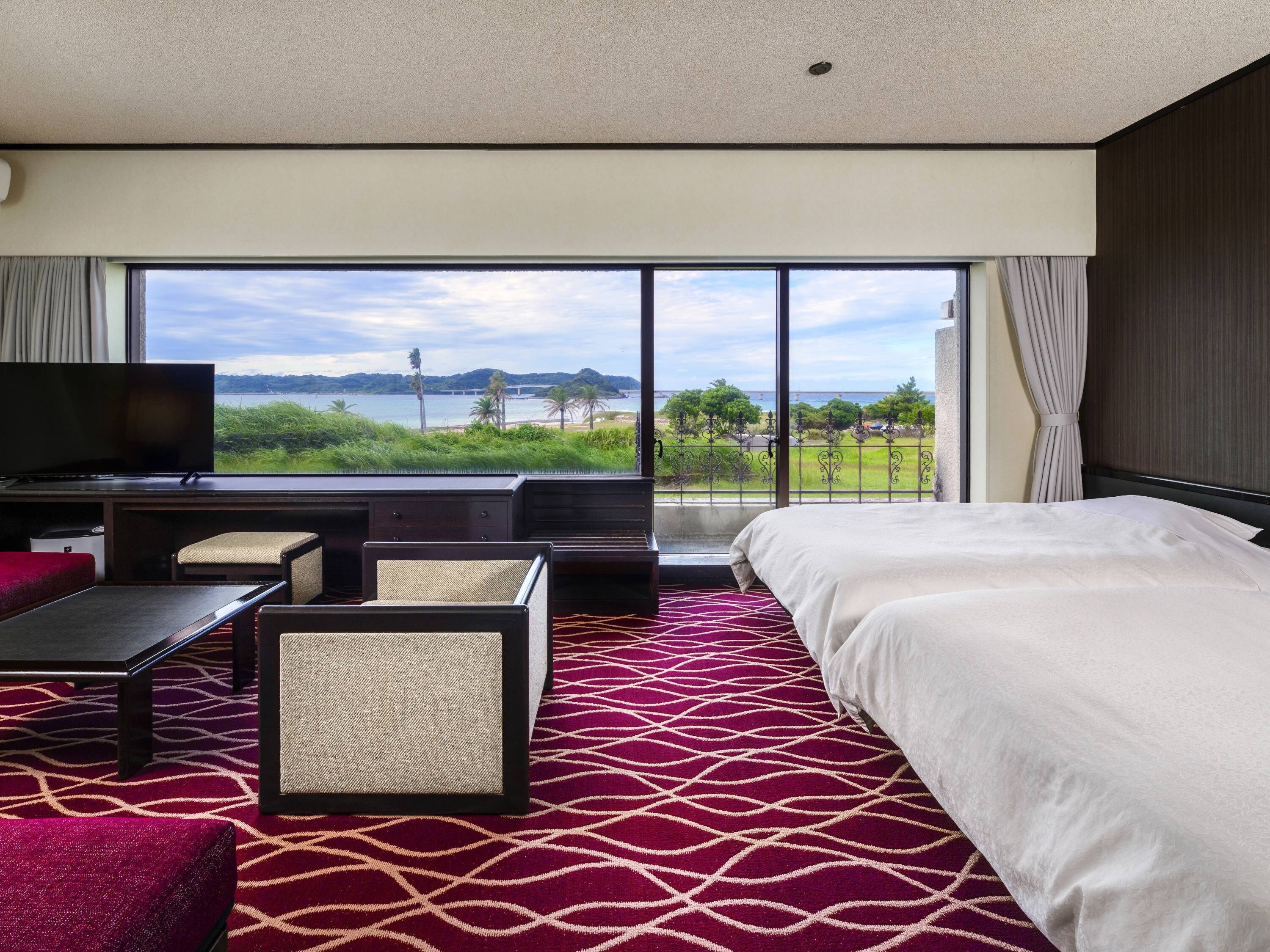 A hotel room with a large bed, a television, a desk, and a view of a body of water and greenery.