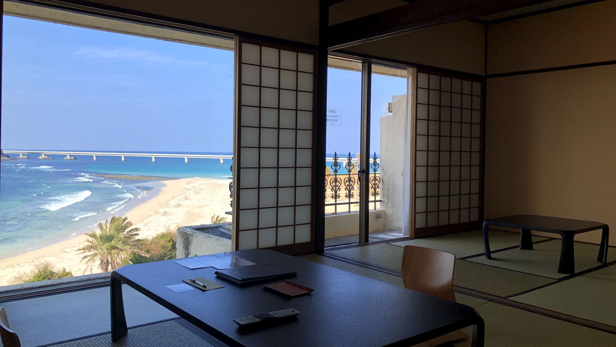 A room with tatami flooring, a table with writing materials, and a view of the beach and ocean.