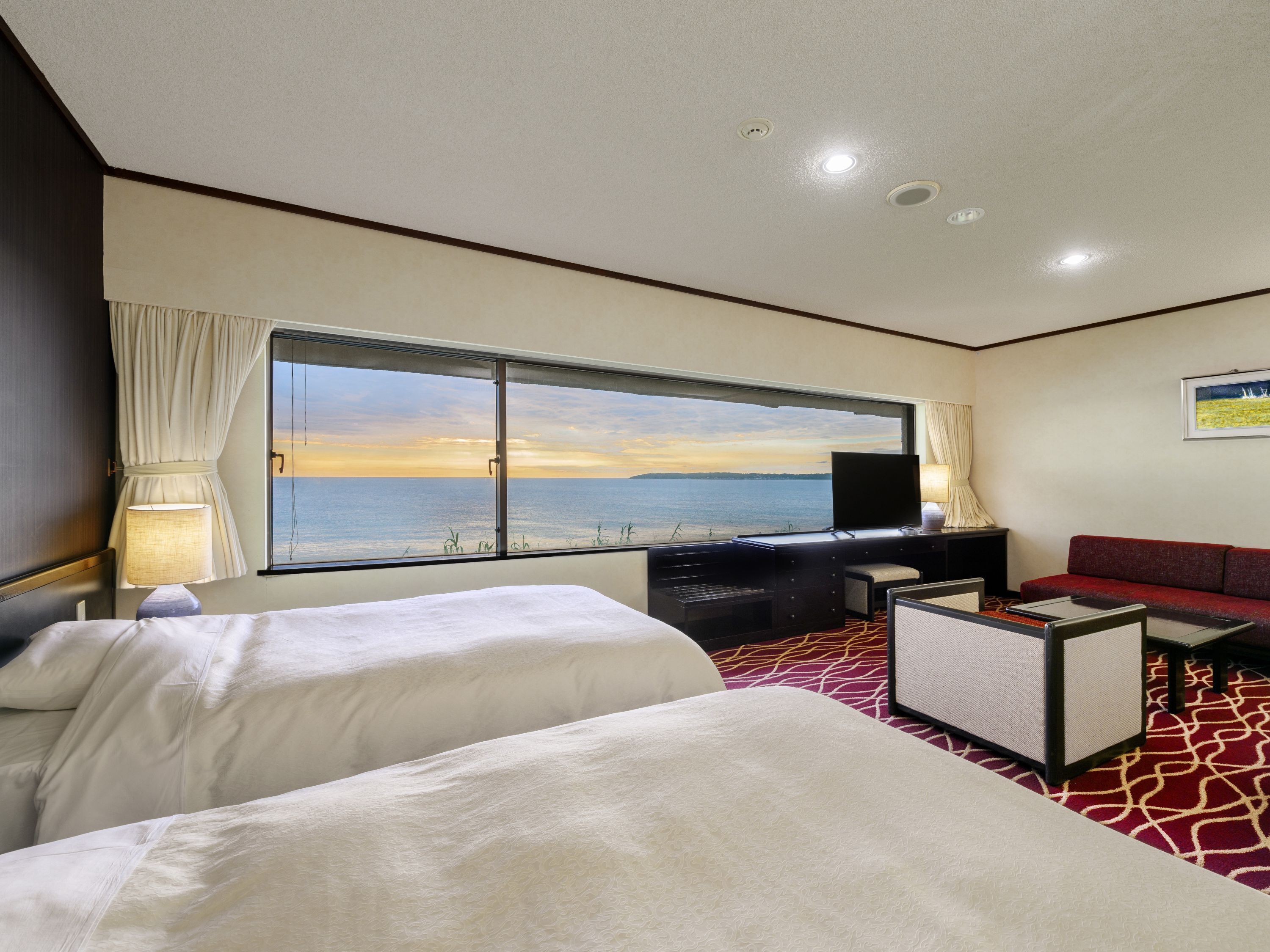 A hotel room with two beds, a desk with a television, a sofa, and a large window with a view of the sea.