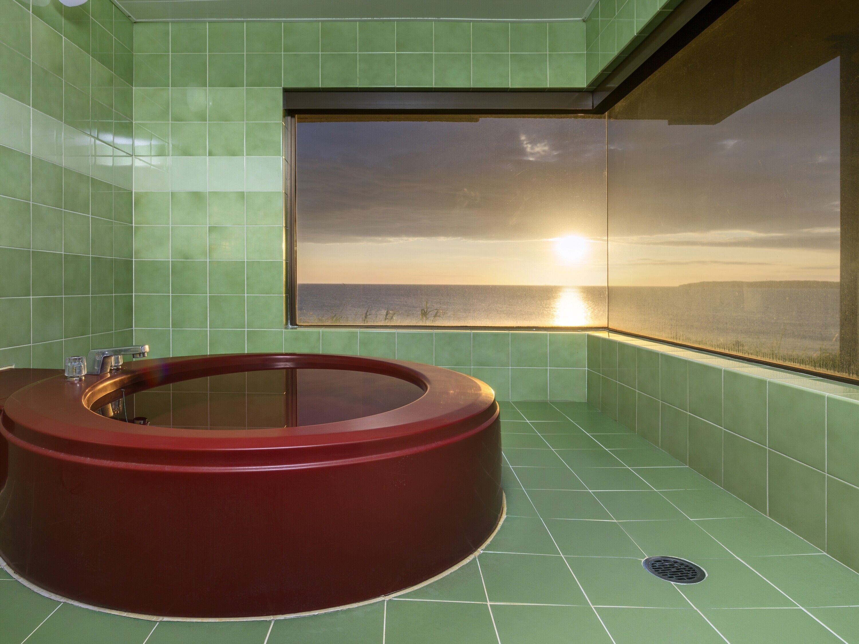 Un baño con una bañera circular, paredes de azulejos verdes y una ventana con vista al atardecer.
