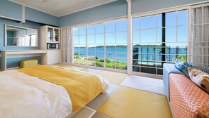 Een hotelkamer met een groot raam met uitzicht op zee, een bed met een gele deken, een bank met decoratieve kussens, en een klein keukentje met een magnetron.