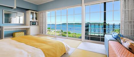 A hotel room with a large window offering a view of the sea, a bed with a yellow blanket, a sofa with patterned cushions, and a small kitchen area with a microwave.
