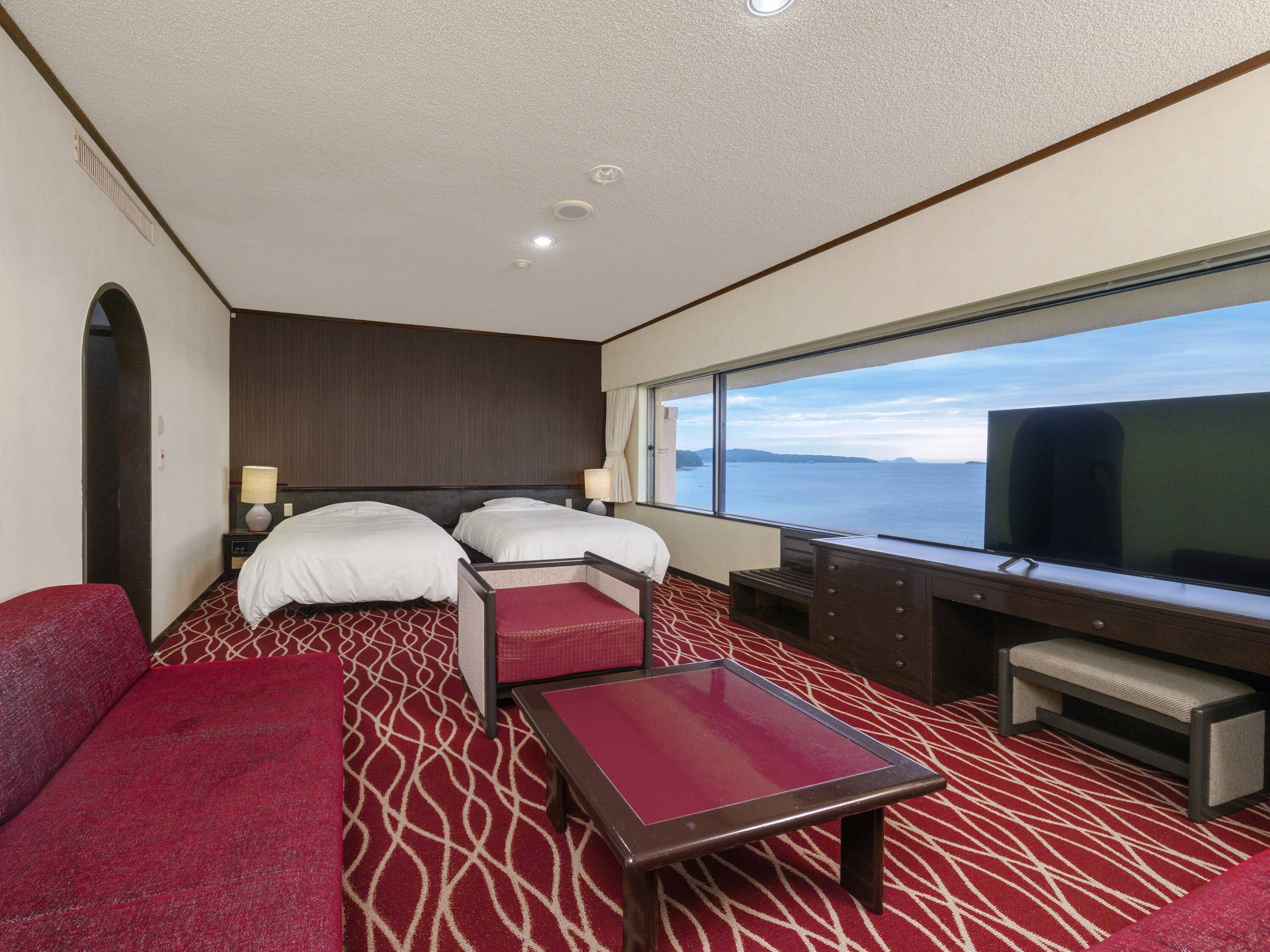 A hotel room with two beds, a desk, a TV, and a large window with a view of the sea.