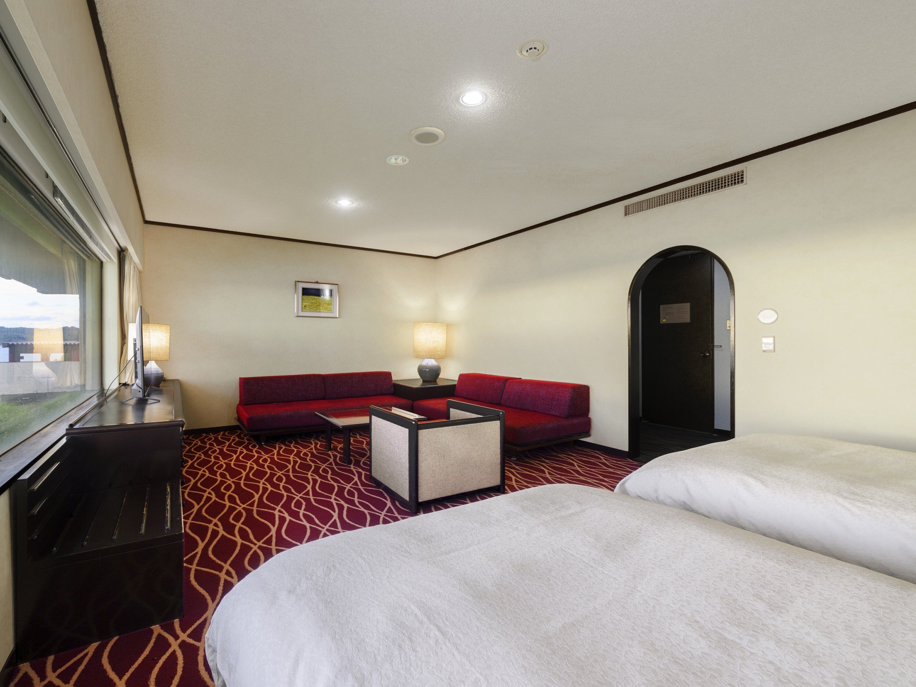 Una habitación de hotel con un sofá rojo, dos camas, un escritorio con lámpara y una ventana con cortinas.