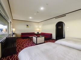 Una habitación de hotel con un sofá rojo, dos camas, un escritorio con lámpara y una ventana con cortinas.