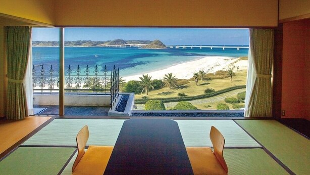 Een kamer met tatami matten, een lage tafel en stoelen. Het heeft een groot raam met uitzicht op een strand, palmbomen en een brug.