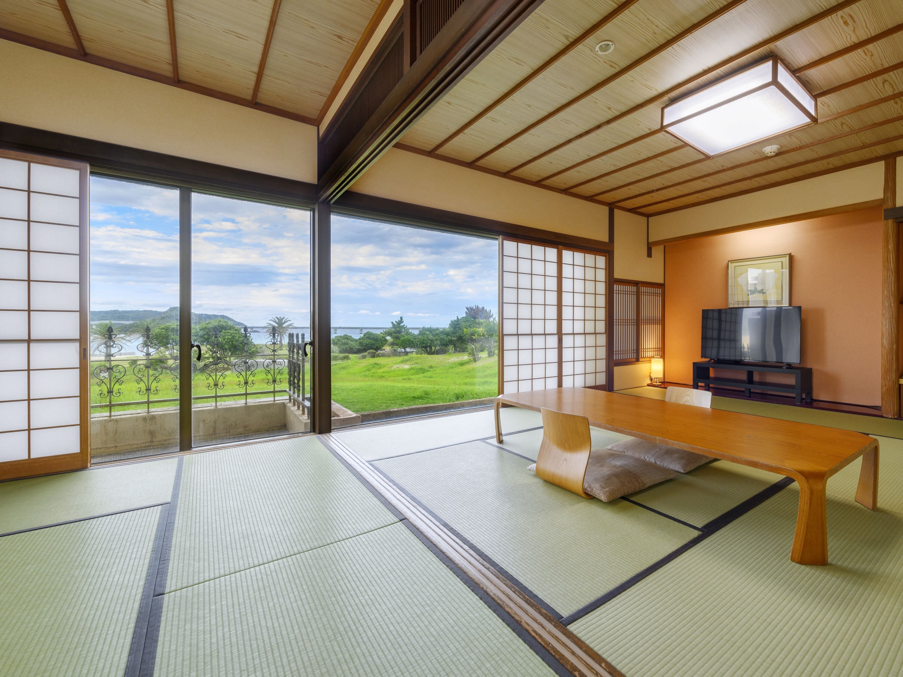 Um quarto japonês tradicional com tatames, uma mesa baixa, uma televisão e vista para uma paisagem verde.