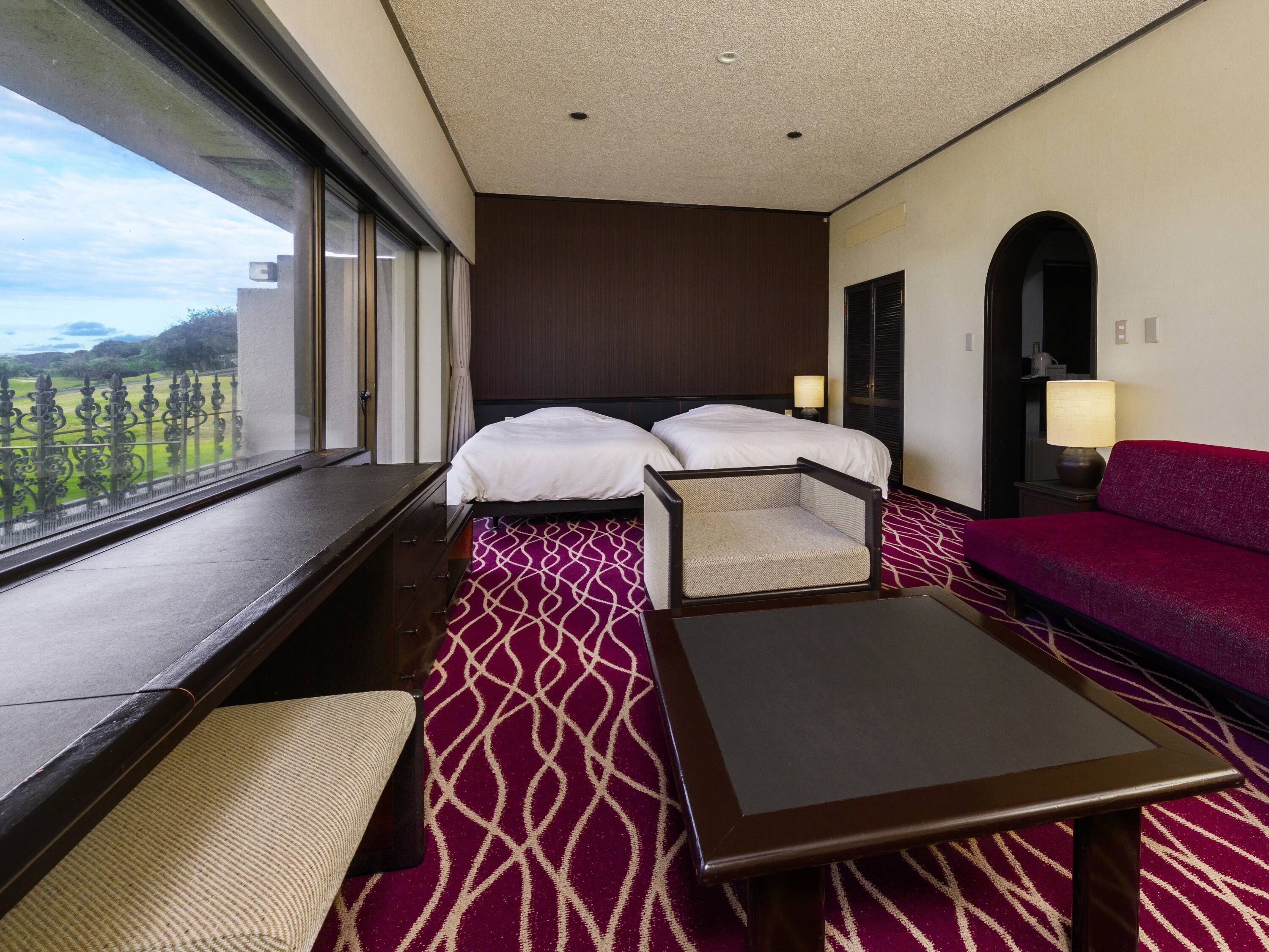 A hotel room with two beds, a sofa, a small table, and a window with a view of a green landscape.