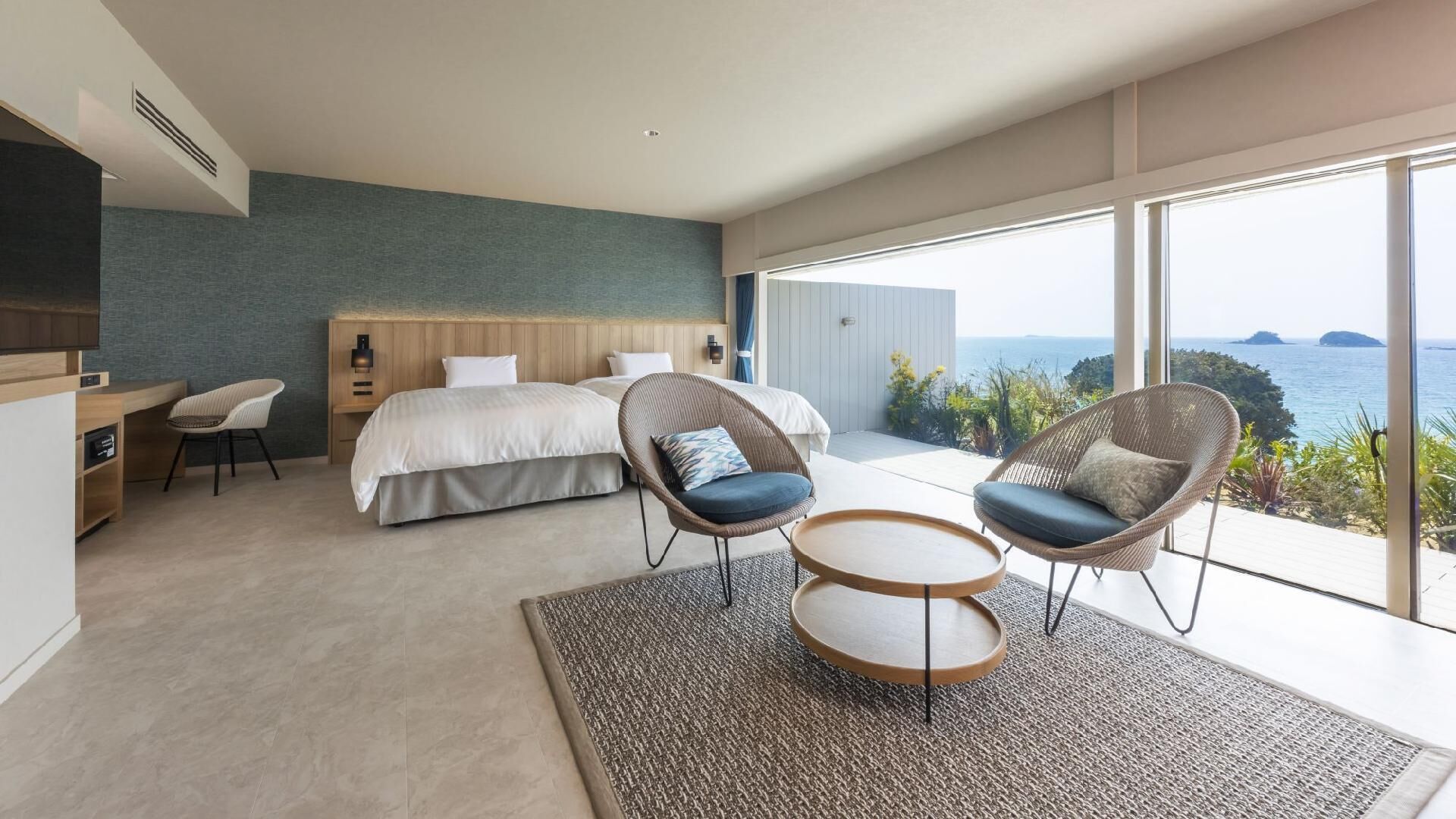 Ein modernes Schlafzimmer mit einem großen Bett, zwei Korbstühlen, einem kleinen Tisch und Meerblick.