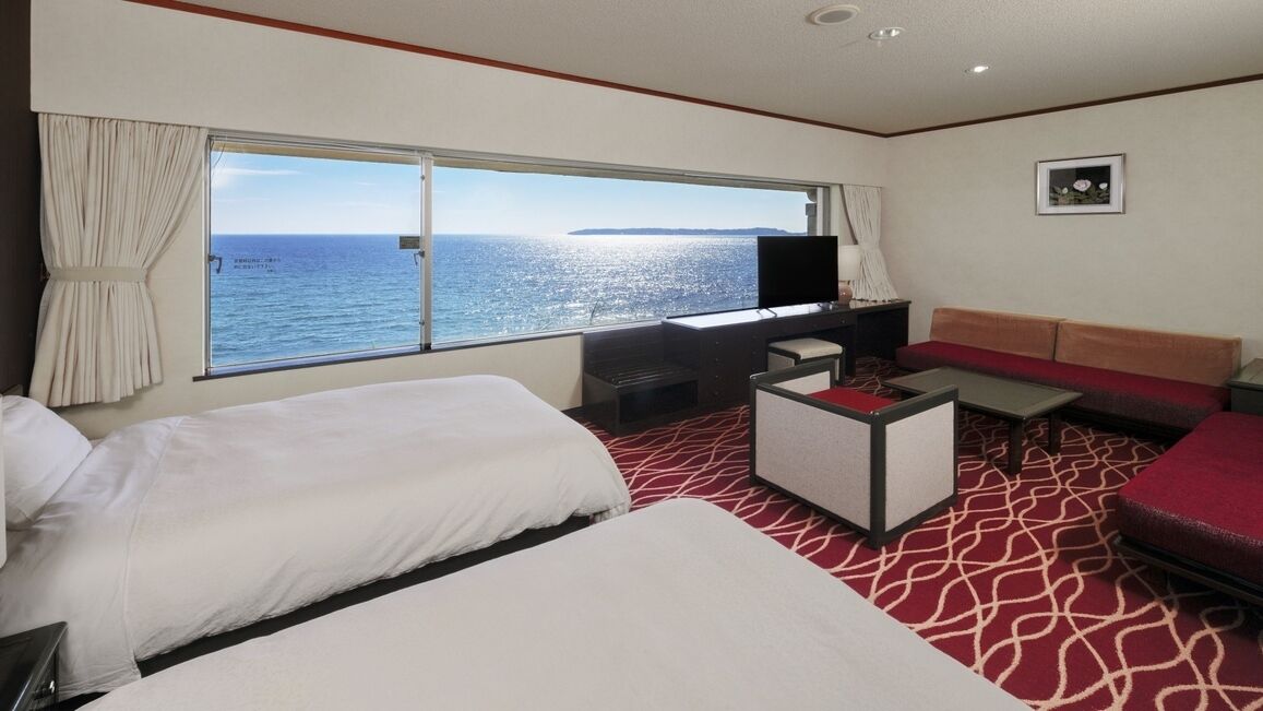 A hotel room with two beds, a desk, a television, a sofa, and a large window with a view of the sea.