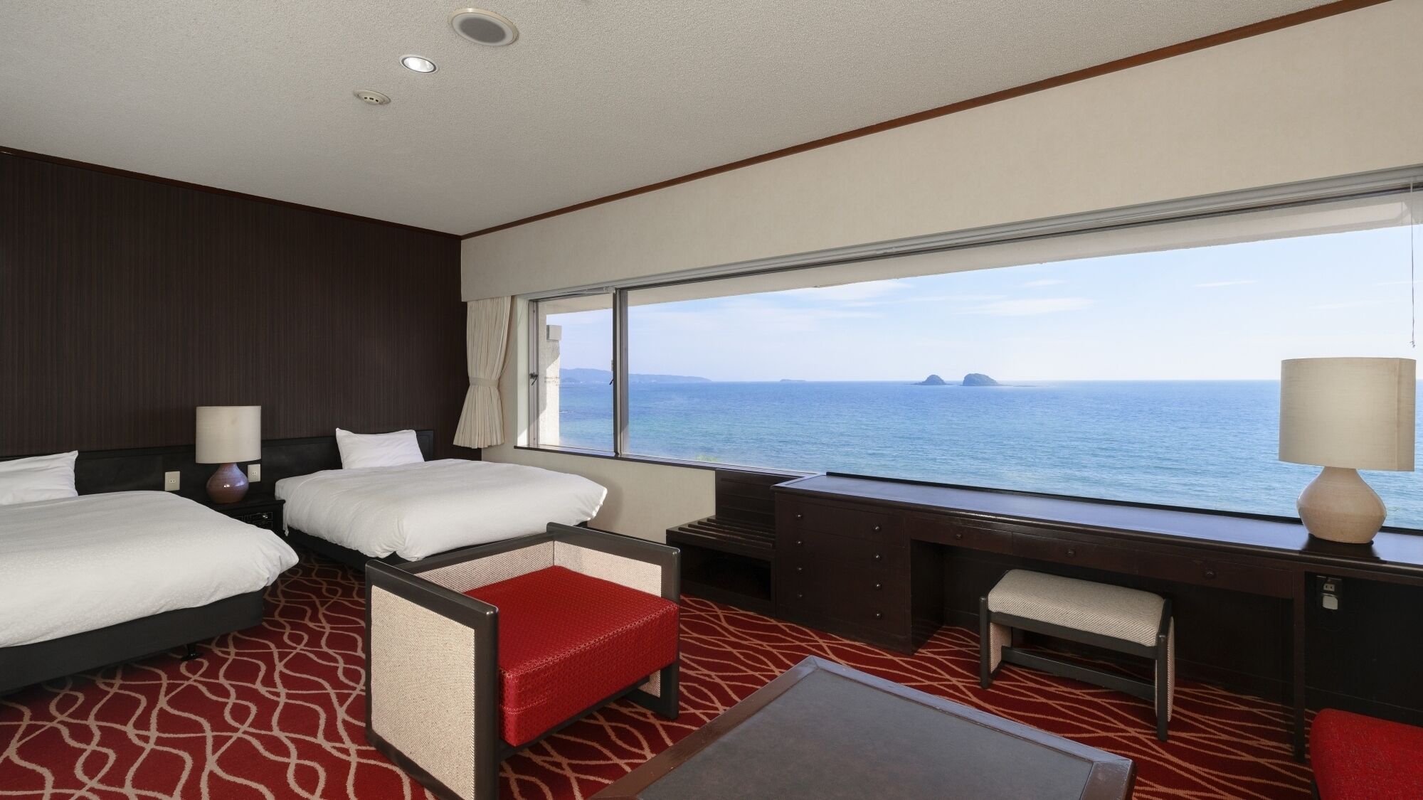 A hotel room with two beds, a desk, a chair, and a view of the sea.