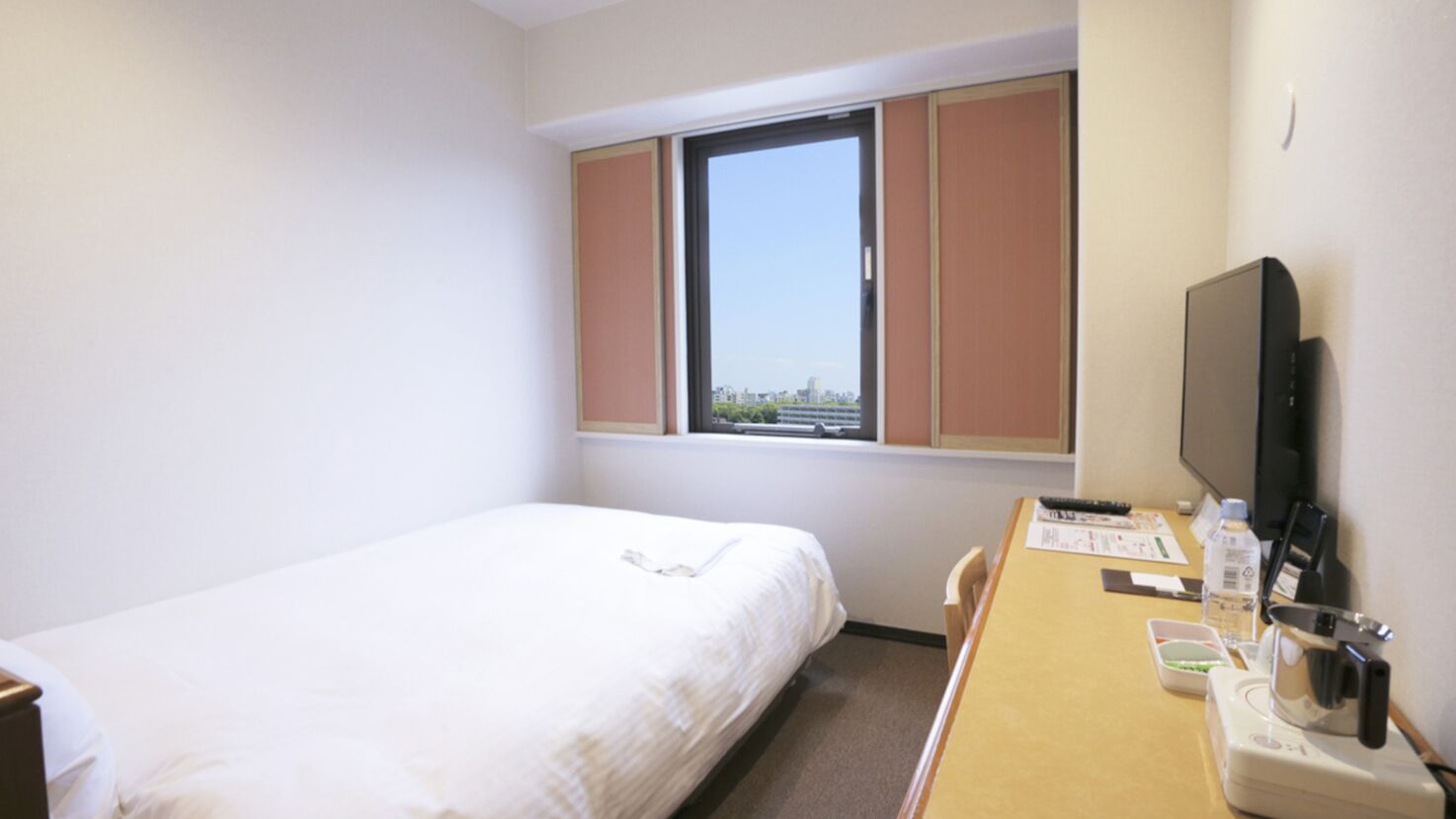 A hotel room with a single bed, a window with blinds, a desk with a TV, and a view of a cityscape.