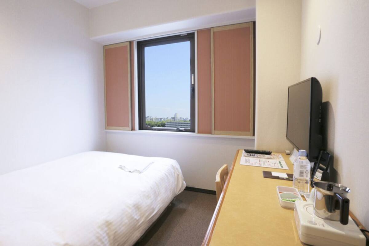 A hotel room with a single bed, a window with blinds, a desk with a TV, and a view of a cityscape.