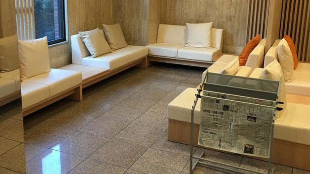 A modern lobby with a seating area featuring white sofas and benches, a tiled floor, and a newspaper rack. - Smile Hotel Sugamo (Tokyo)