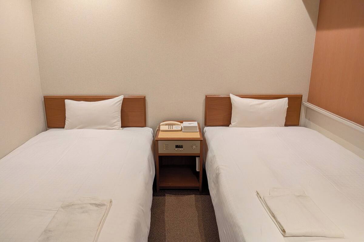 A hotel room with two single beds, a bedside table with a telephone and a book, and a carpeted floor.