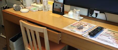 A hotel room desk with a computer, a chair, a trash bin, and a small refrigerator.