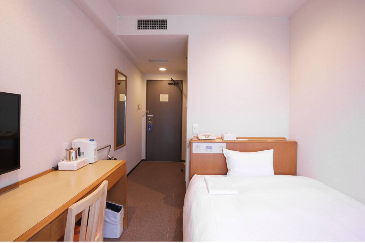 A hotel room with a bed, bedside table, a desk with a chair, a mirror, a television, and a door leading to another room.