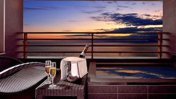 Uma varanda com vista para o pôr do sol sobre o mar, uma garrafa de champanhe e duas taças sobre uma mesa.
