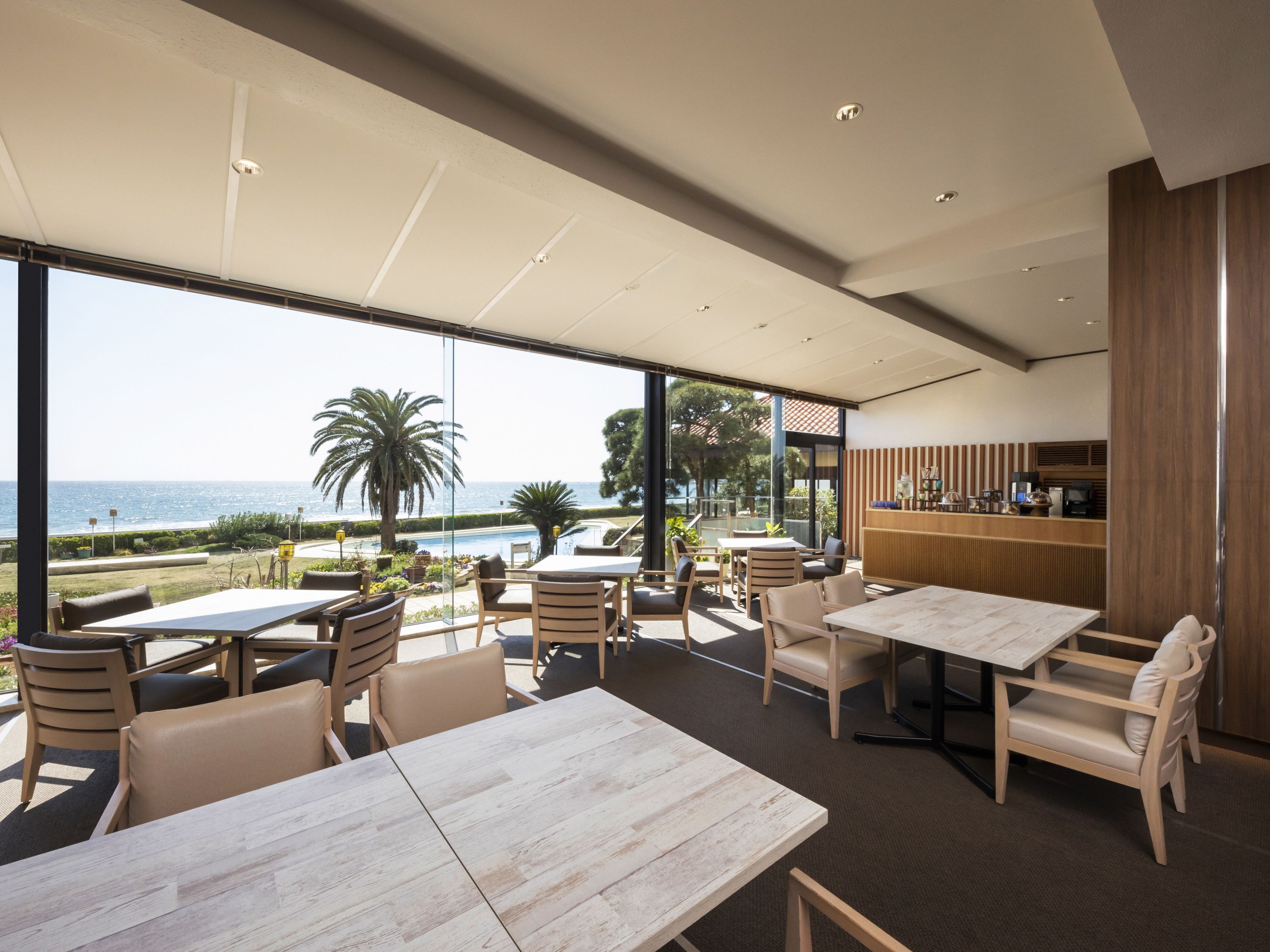 A modern outdoor dining area with tables and chairs, overlooking a pool and a view of the sea.