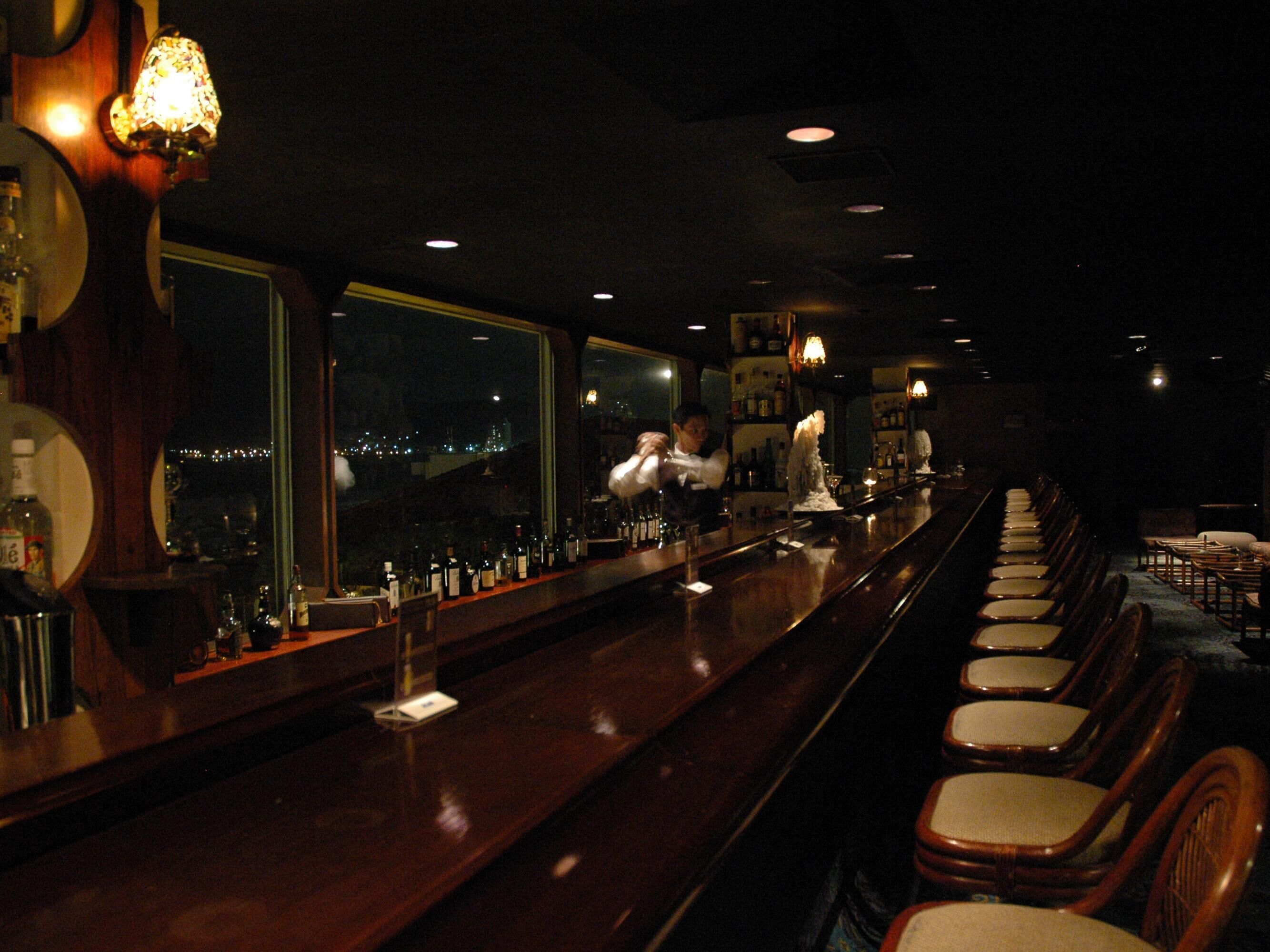 Een bar met een lange toonbank, diverse flessen en een barman.
