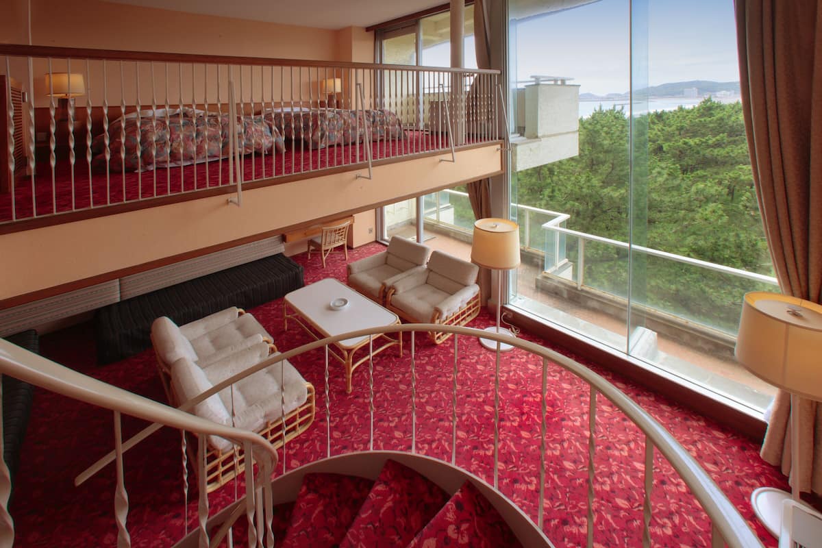 Hotelkamer met een roodgekleurd tapijt, een balkon met een leuning, een bank, een bijzettafeltje en een lamp.