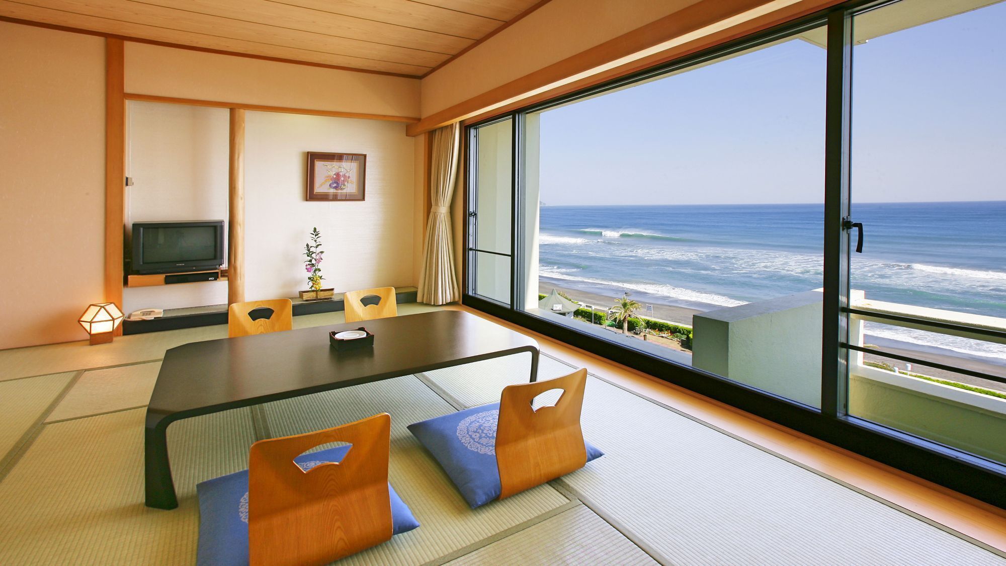 A room with tatami flooring, a low table, chairs, a television, and a large window with a view of the ocean.