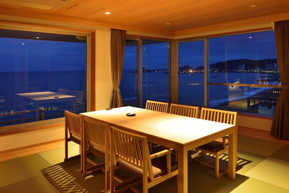 Een eetruimte met een tafel en stoelen, grote ramen met uitzicht op zee en stadslichten, en een houten plafond.