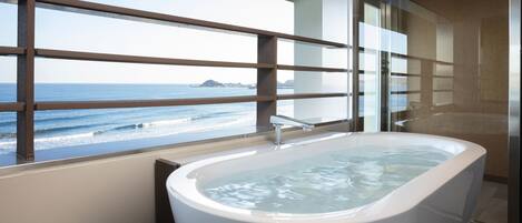 A modern bathroom with a freestanding bathtub, large windows with a view of the ocean, and a wooden balcony railing.