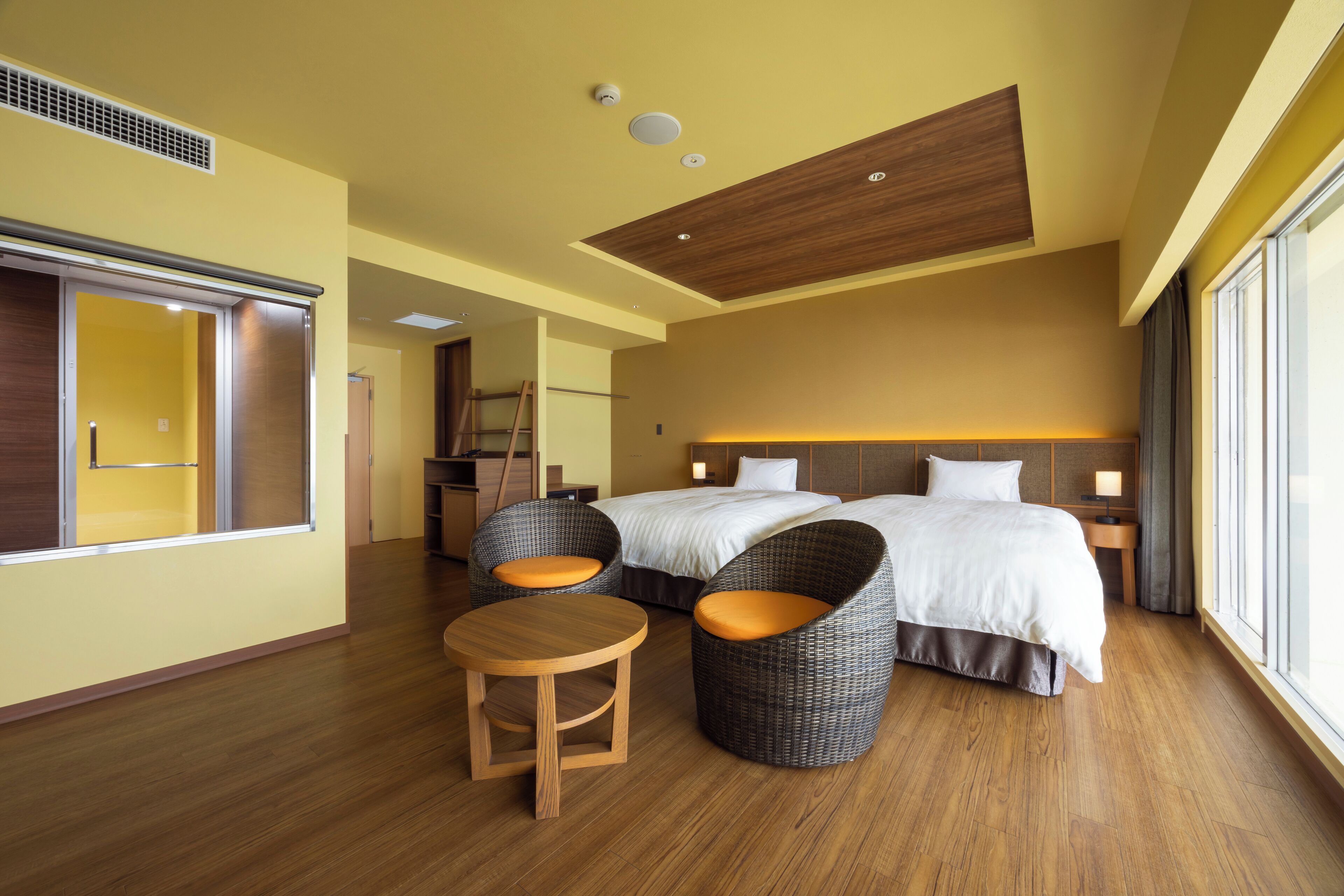 Une chambre d’hôtel avec un grand lit, deux fauteuils en osier, une petite table en bois et une grande fenêtre.
