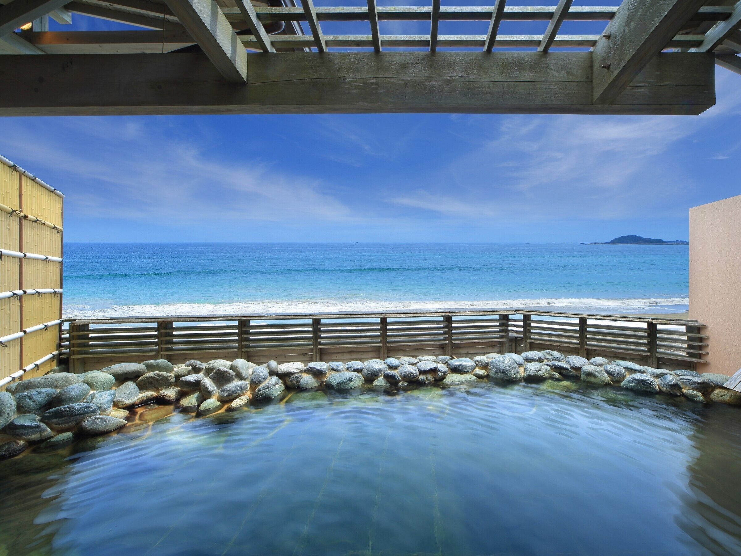 A stone-lined pool with a wooden deck and a view of the beach and ocean.