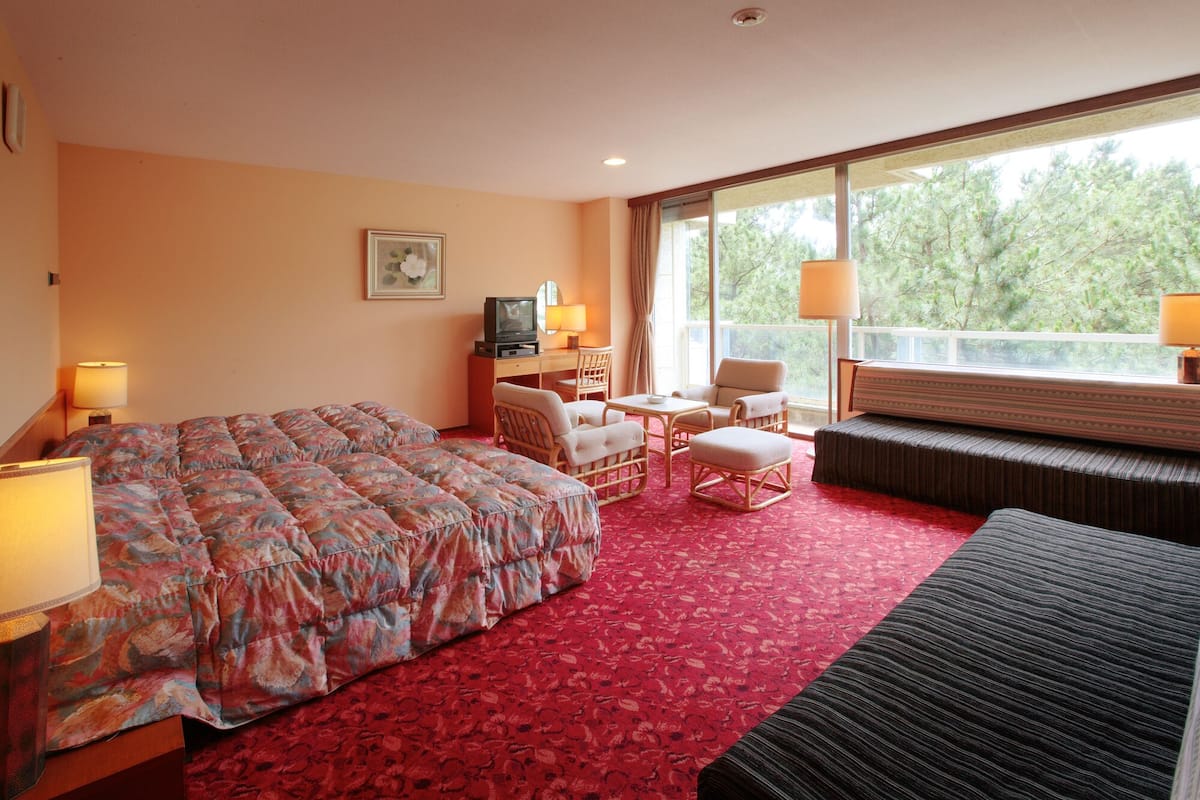 Een hotelkamer met een bed, een bureau met een televisie, een stoel, een bank en een groot raam met uitzicht op bomen.