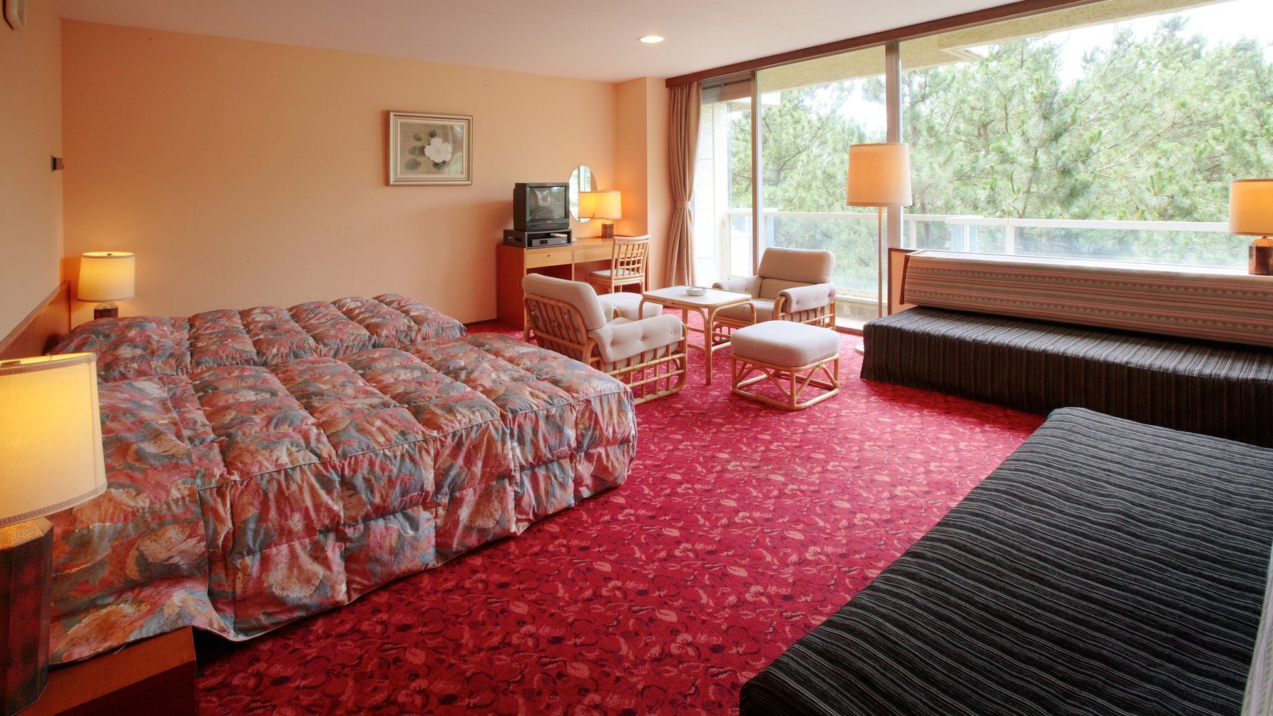 A hotel room with a large bed, a sofa, two chairs, a small table, a TV, and a window with a view of trees.