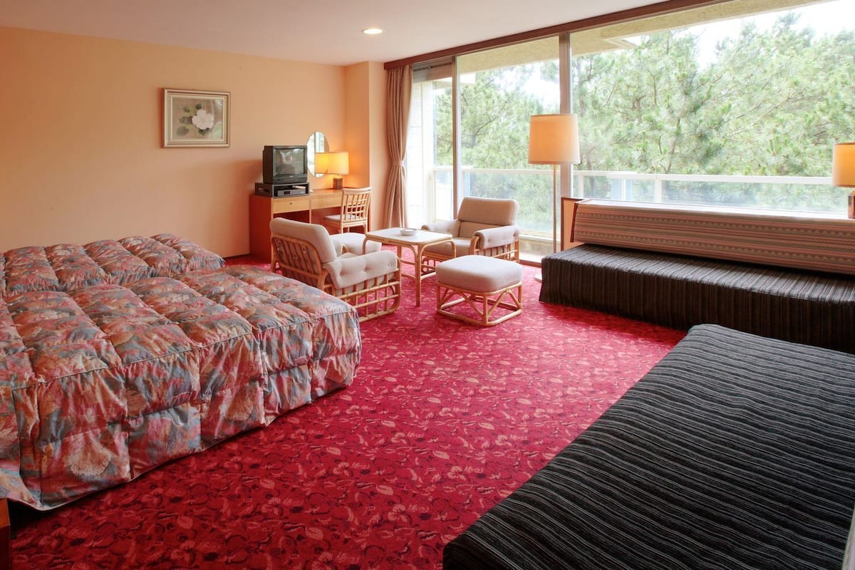 Une chambre d’hôtel avec un grand lit, un canapé, deux fauteuils, une petite table, un téléviseur et une fenêtre donnant sur des arbres.