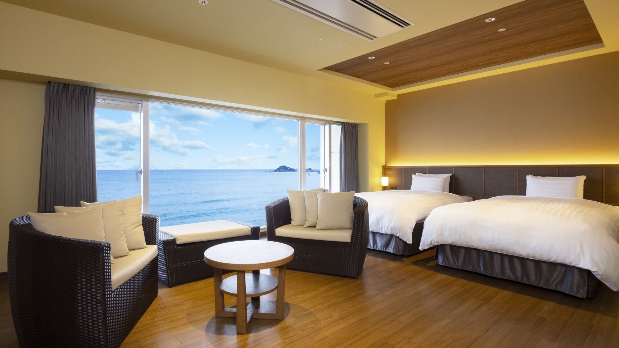 Habitación de hotel con dos camas, un sofá, una mesa redonda y un ventanal con vistas al mar.
