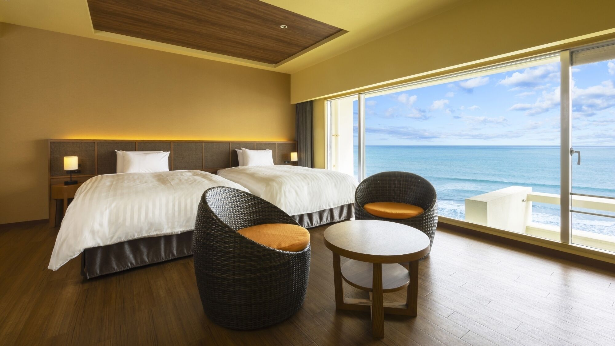 Habitación de hotel con dos camas, una silla de mimbre, una mesita redonda y un ventanal con vista al océano.