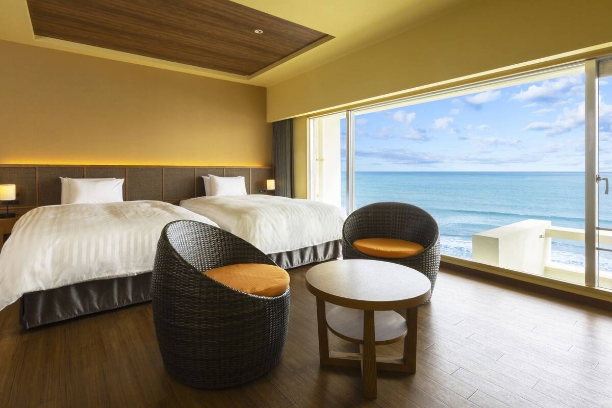 Een hotelkamer met twee bedden, een rieten stoel, een klein rond tafeltje en een groot raam met uitzicht op de oceaan.